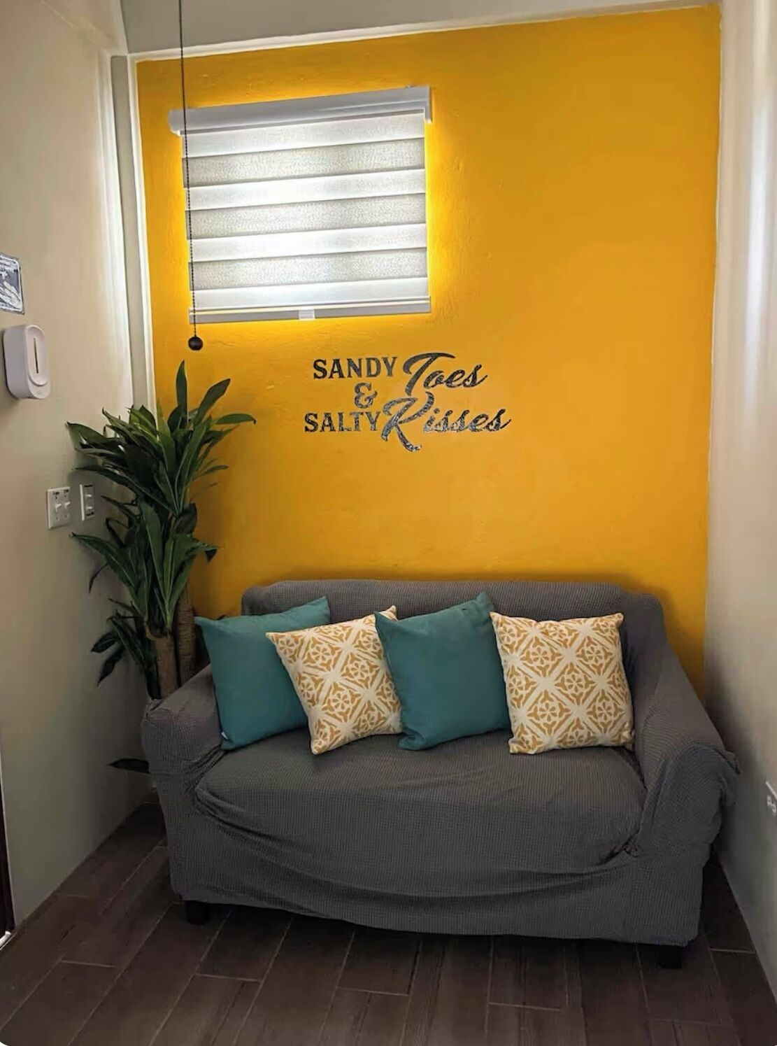 Una habitación con sofá gris, pared amarilla y ventana con persianas.