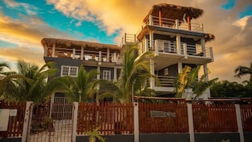 Un edificio de varios pisos con techo de paja, una entrada con reja y palmeras.