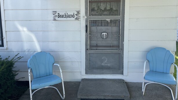 Et hvidt hus med en dør, der står "Beachland" på, og to blå stole foran.