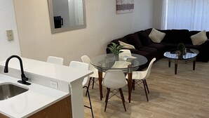 A modern kitchen with a white countertop and wooden cabinets, a dining area with a glass table and chairs, and a living room with a dark sofa and a window with curtains.