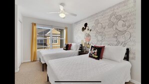 A hotel room with two beds, a ceiling fan, a window with yellow curtains, and a wall with Mickey Mouse wallpaper.