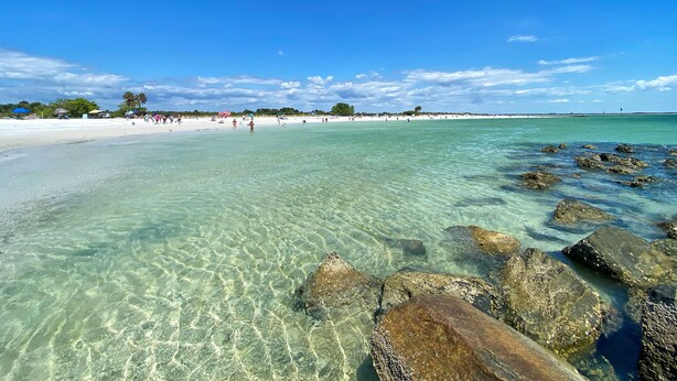 A beach with clear shallow waters, rocks in the foreground, and a sandy shore with people in the distance. - Spacious & Fun Family Gem / Pool, Beach, Blue Jays (Palm Harbor)