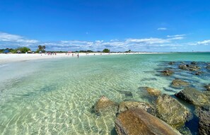 Una playa con aguas cristalinas y poco profundas, rocas en primer plano y una orilla de arena con gente a lo lejos.