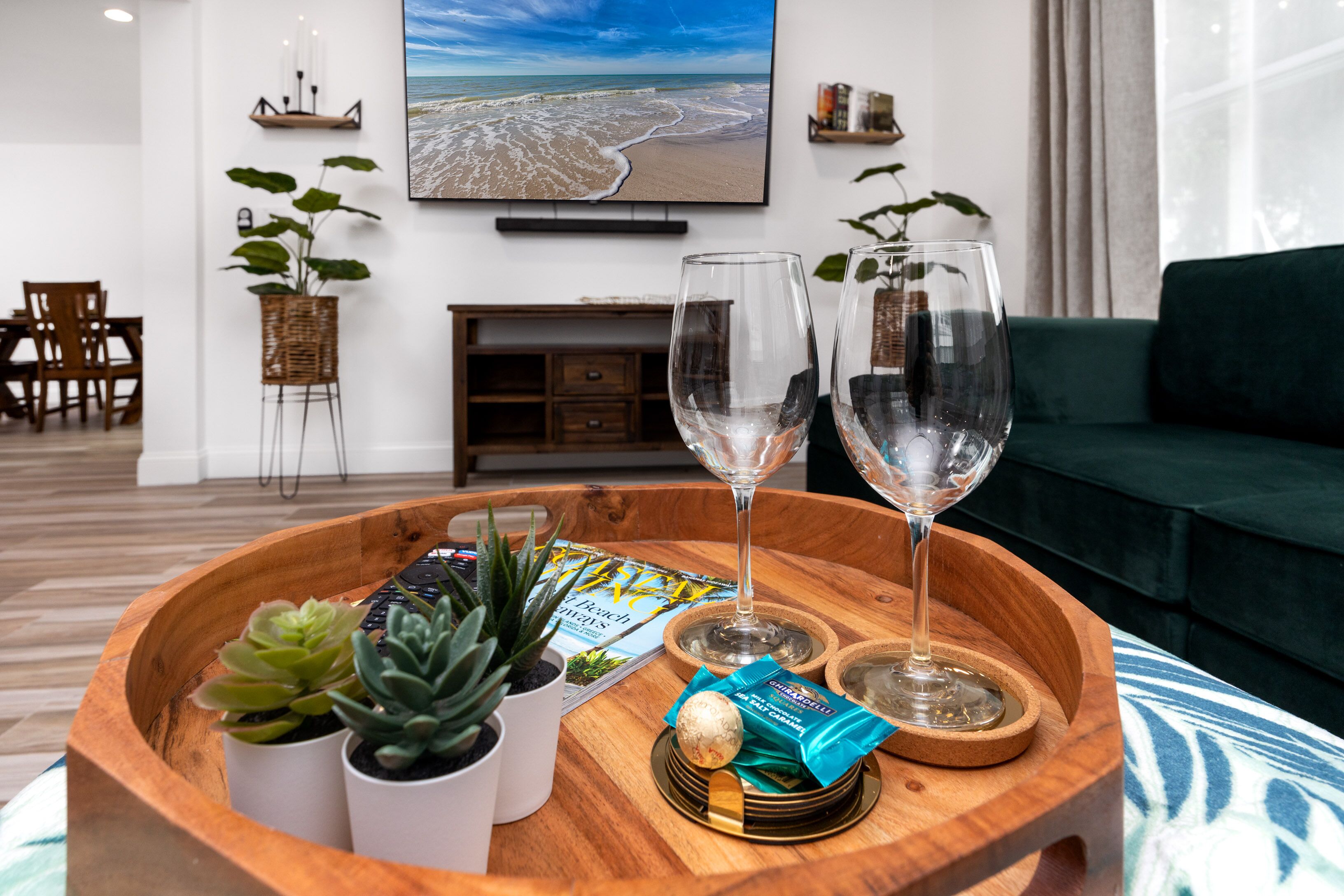 Una bandeja de madera con dos copas de vino, una planta suculenta, una planta pequeña en maceta y una revista sobre una mesa en un moderno salón con un sofá verde y un televisor que muestra una escena de playa.