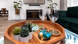 A wooden tray with two wine glasses, a succulent plant, a small potted plant, and a magazine on a table in a modern living room with a green sofa and a TV displaying a beach scene. - Spacious & Fun Family Gem / Pool, Beach, Blue Jays (Palm Harbor)