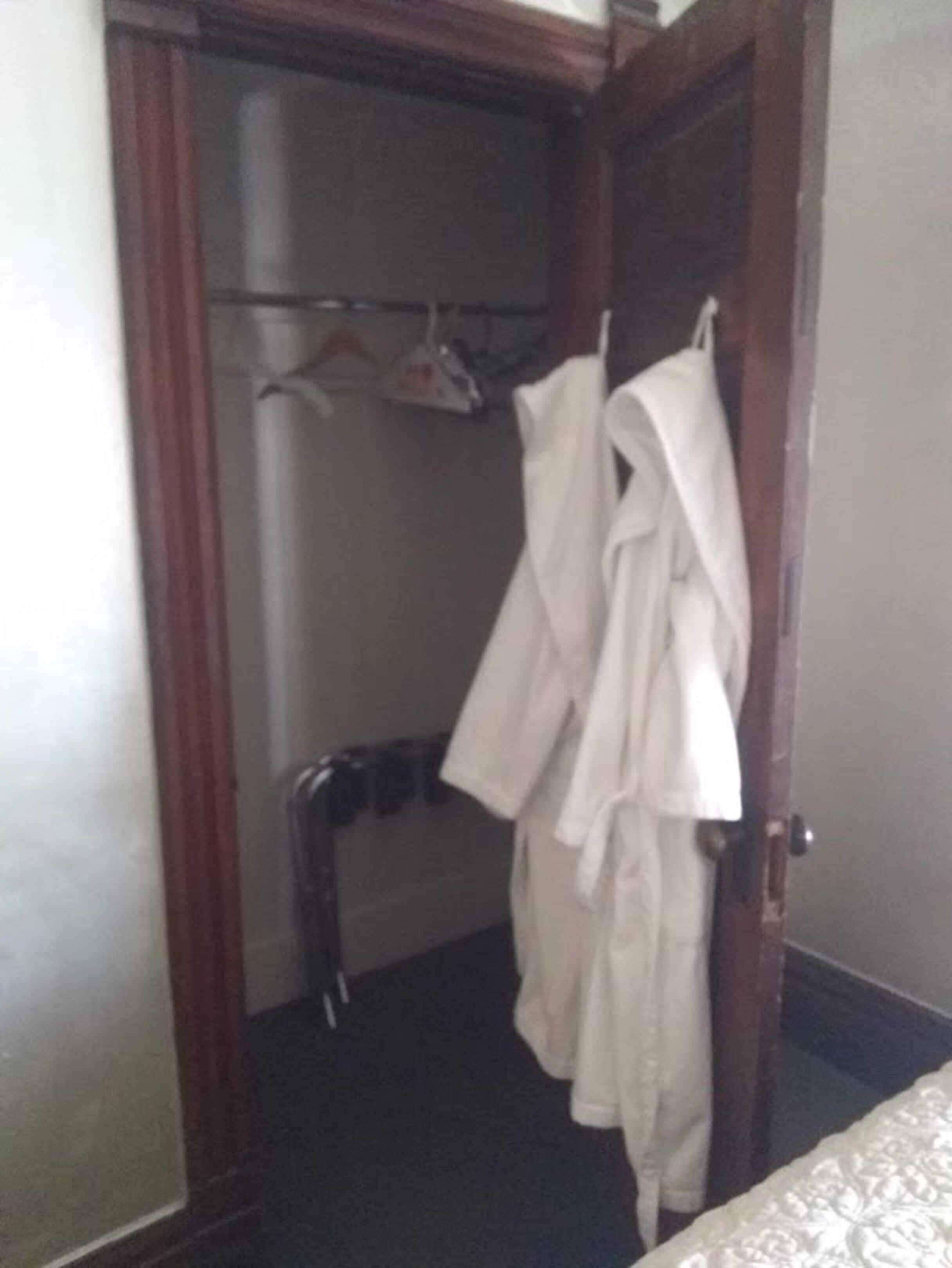 A hotel room with a wooden door, a closet with hanging robes, and a bed with a white bedspread.