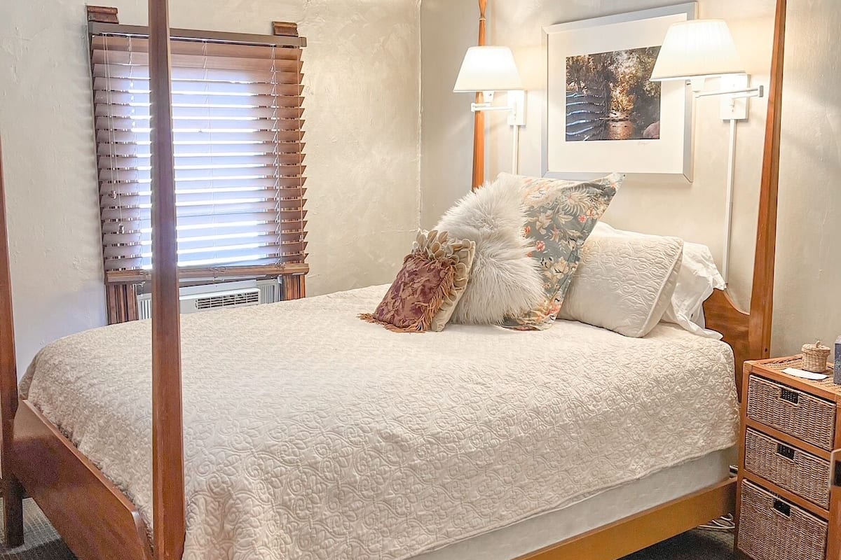 A four-poster bed with a white bedspread, two wall-mounted lamps, and a framed picture on the wall.