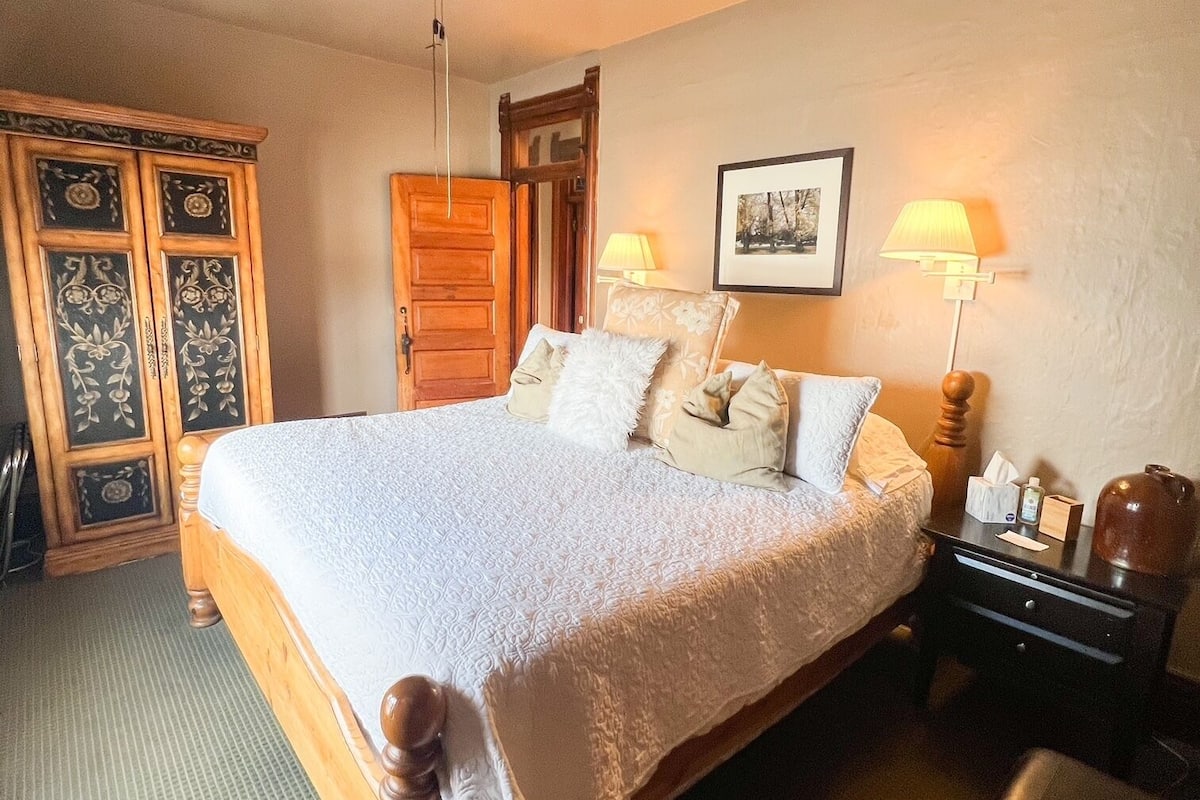 A neatly arranged bedroom with a large bed, a ceiling fan, a wooden wardrobe, and a bedside table with a lamp.