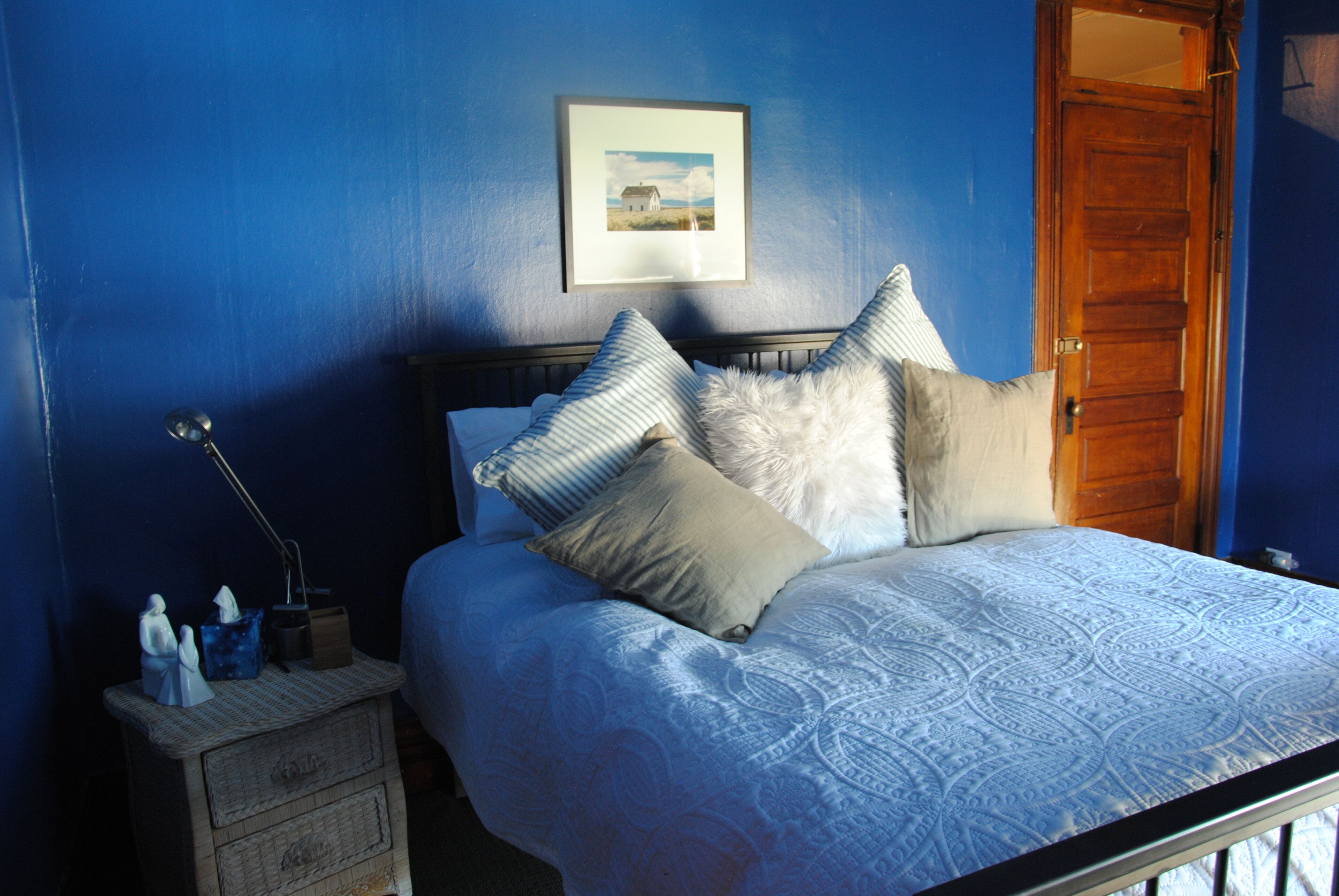 Une chambre à coucher avec un lit, des oreillers, une table de chevet, une lampe, un tableau encadré au mur et une porte en bois.