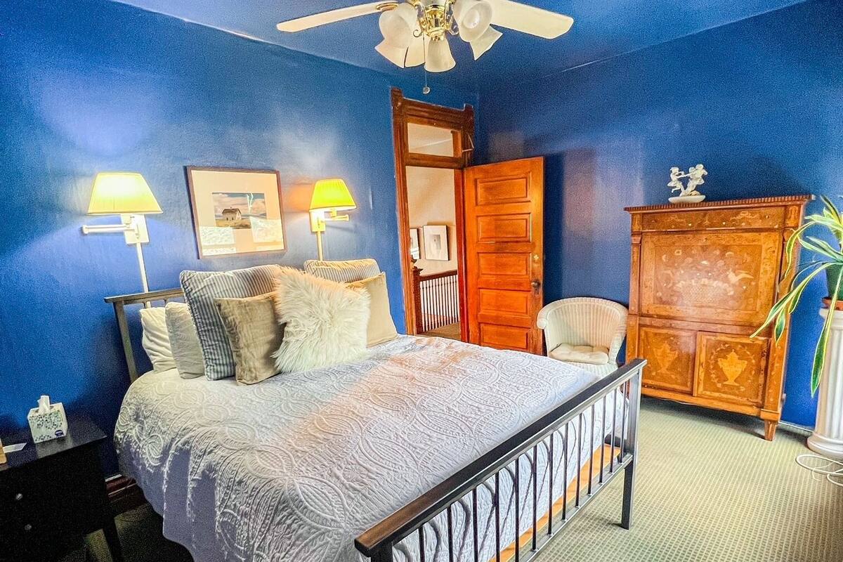 A bedroom with a bed, two bedside lamps, a ceiling fan, a wooden wardrobe, and a door.