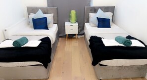 Een kamer met twee bedden, elk met een grijze hoofdbord, witte beddengoed en teal- en zwartgekleurde dekens. Tussen de bedden staat een witte nachtkastje met een groene vaas.