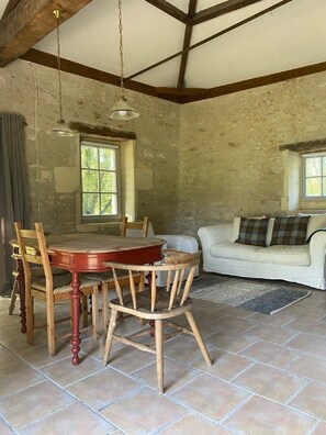 A rustic room with exposed wooden beams, a round wooden table with chairs, a white sofa, and two windows with curtains. - Beautiful Boathouse by the River (Bourg-Charente)