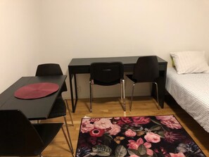 A small, tidy room with a desk, two chairs, a bed with a white pillow, and a floral rug. - Apartment in Årsta Stockholm 237 (Arsta)