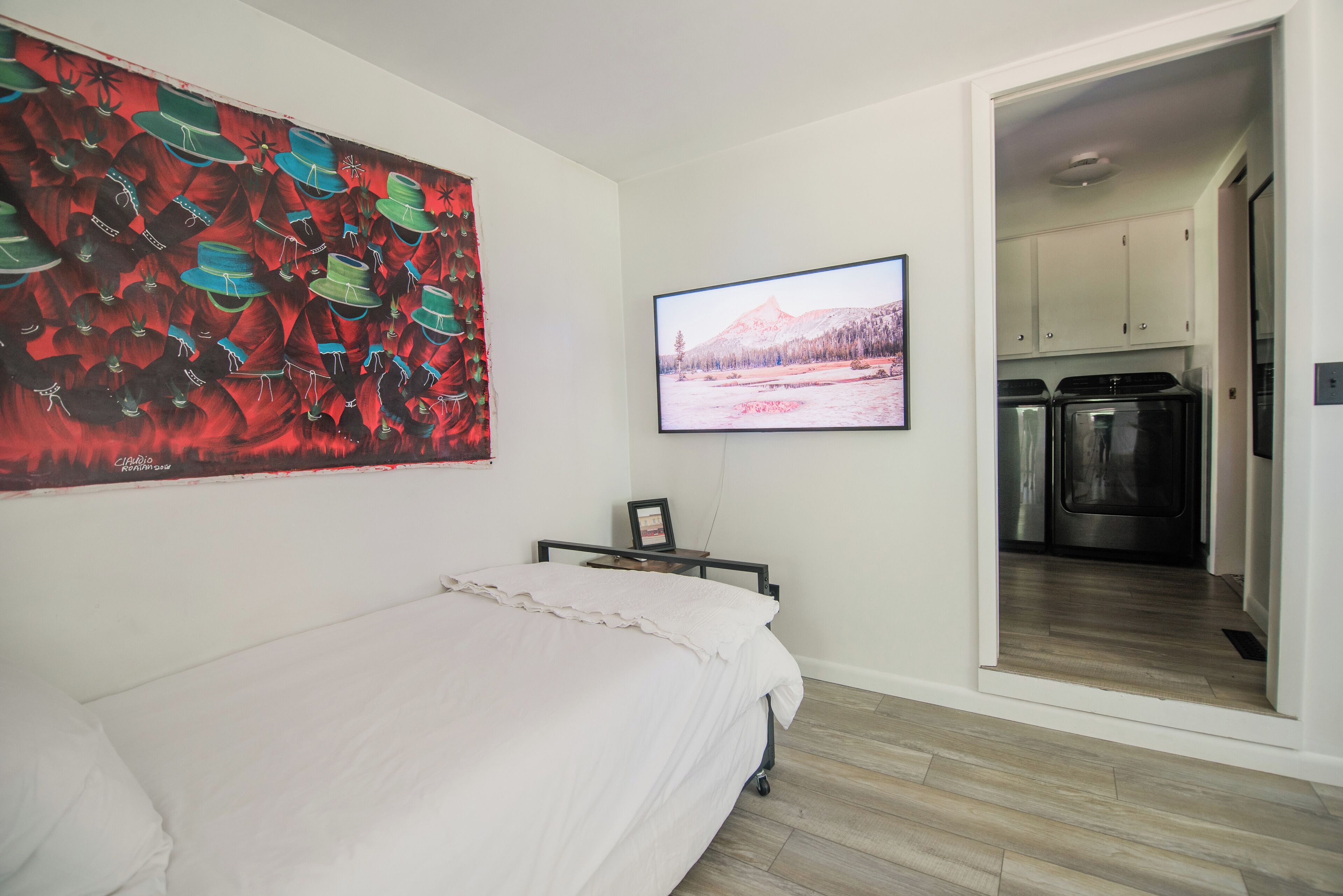 A bedroom with a bed, a painting, a television, and a view into the kitchen.
