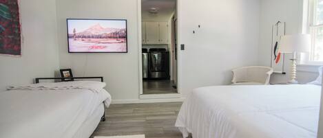 Une chambre avec deux lits, un téléviseur fixé au mur, une cuisine visible à travers une porte ouverte et une lampe blanche sur le côté droit.