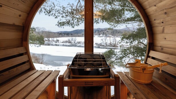 Une cabane en bois dotée d’une grande fenêtre circulaire, d’un barbecue de la marque Harvia et d’un seau en bois.