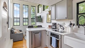 Une cuisine moderne avec des meubles blancs, un lave-vaisselle, un four et une machine à café. Un canapé gris se trouve dans le salon attenant.