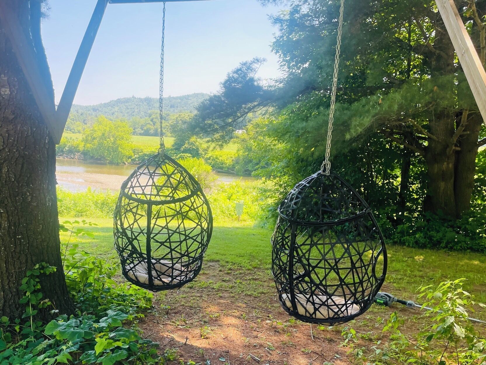 Twee hangende rieten stoelen, bevestigd aan een houten constructie, met uitzicht op een weelderig groen landschap en een rivier in de verte.