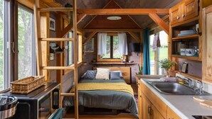 Interior acogedor de una cabaña con una cama, armarios de madera, una zona de cocina y una ventana con vistas a la vegetación.