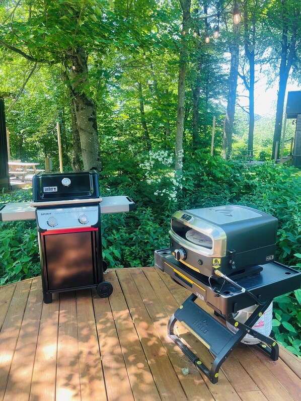 Two outdoor grills, a Weber Spirit and a HALO, on a wooden deck surrounded by greenery. - Maple Sugar Shack | Hot Tub, Sauna, River & Views (Johnson)