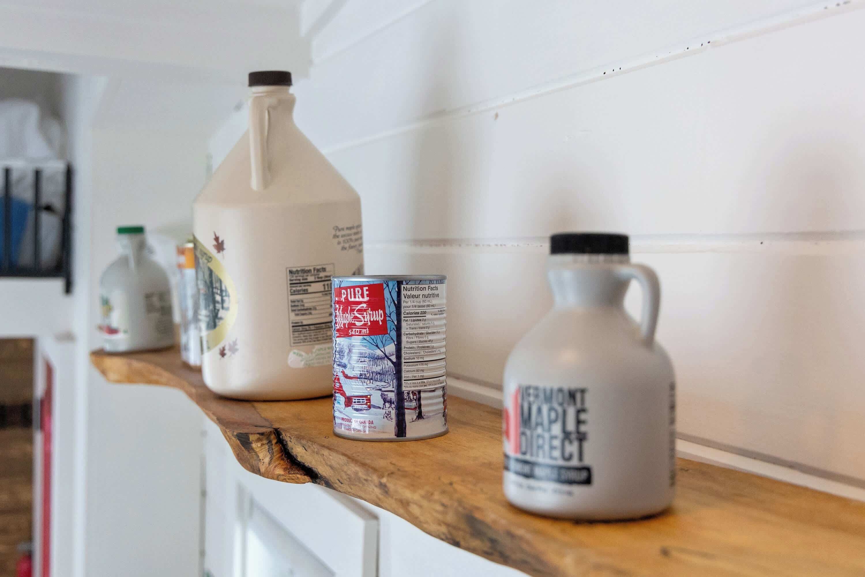 Un plan de travail de cuisine avec une grande cruche blanche, une boîte de sirop d’érable et une plus petite cruche portant l’étiquette « Sirop d’érable – Vente directe ».