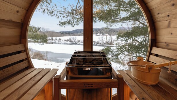 Una cabaña de madera con un ventanal circular grande, un horno de leña Harvia y un cubo de madera.