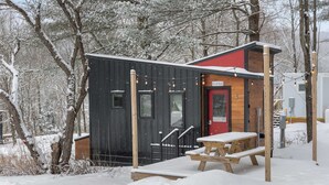 Una cabaña de madera con una puerta roja y una mesa de picnic en un bosque nevado.