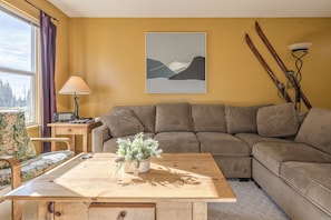 Un salon avec un canapé marron, une table basse en bois ornée d’une plante, une lampe et un tableau abstrait encadré accroché au mur.