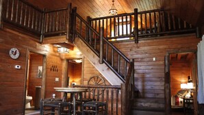 A wooden interior with a staircase, a dining area with chairs and a table, and a visible clock on the wall.
