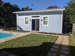 Une maison de plain-pied avec une porte d’entrée, deux fenêtres et un petit jardin à l’avant, avec un chemin en béton menant à une piscine.