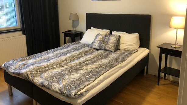 Ein ordentlich bezogenes Bett mit einem gemusterten Überwurf, zwei Nachttischlampen, ein Stadtbild und ein Fenster mit Vorhängen.
