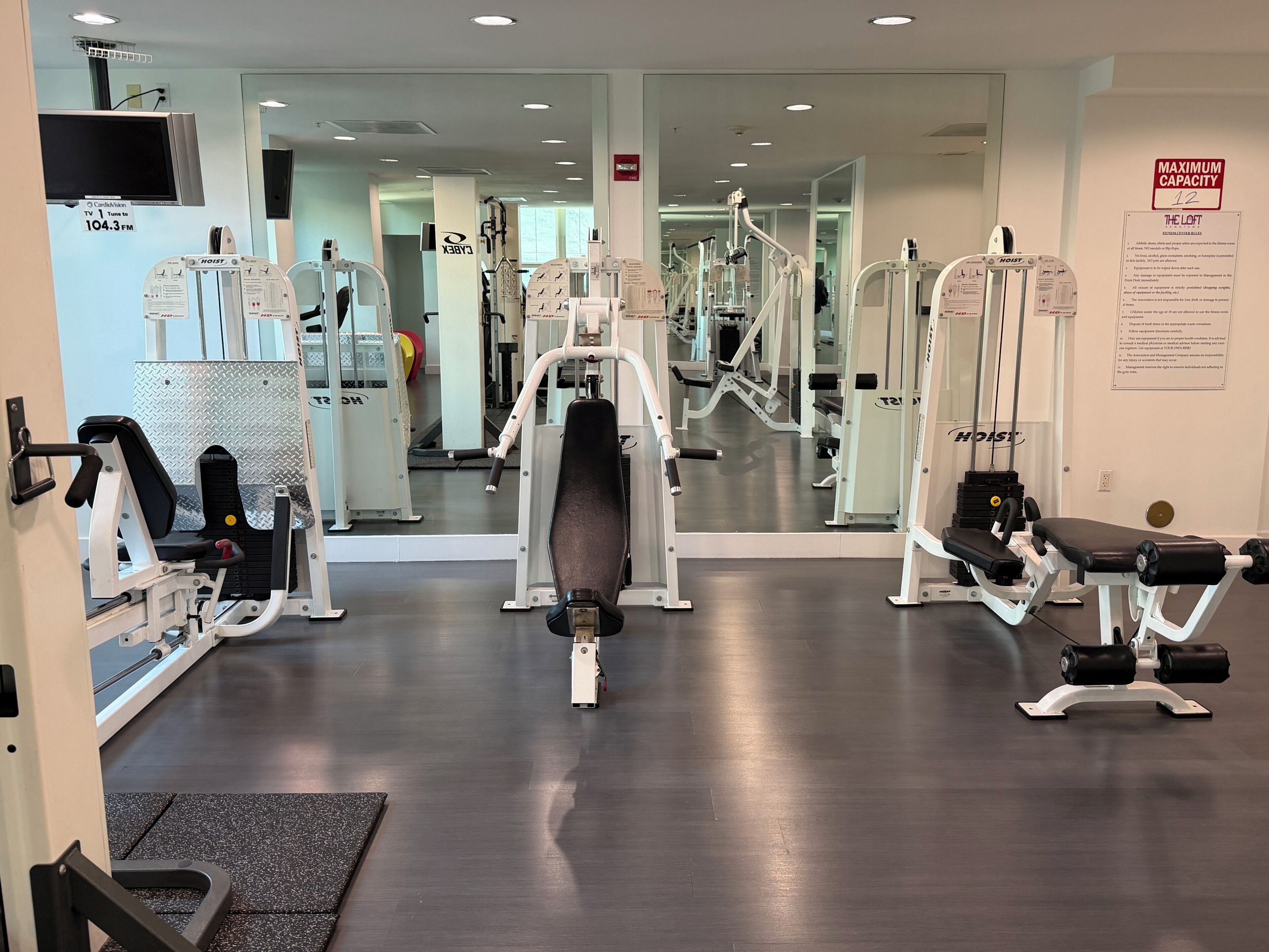 A gym with various exercise machines, a TV mounted on the wall, and a sign indicating maximum capacity.