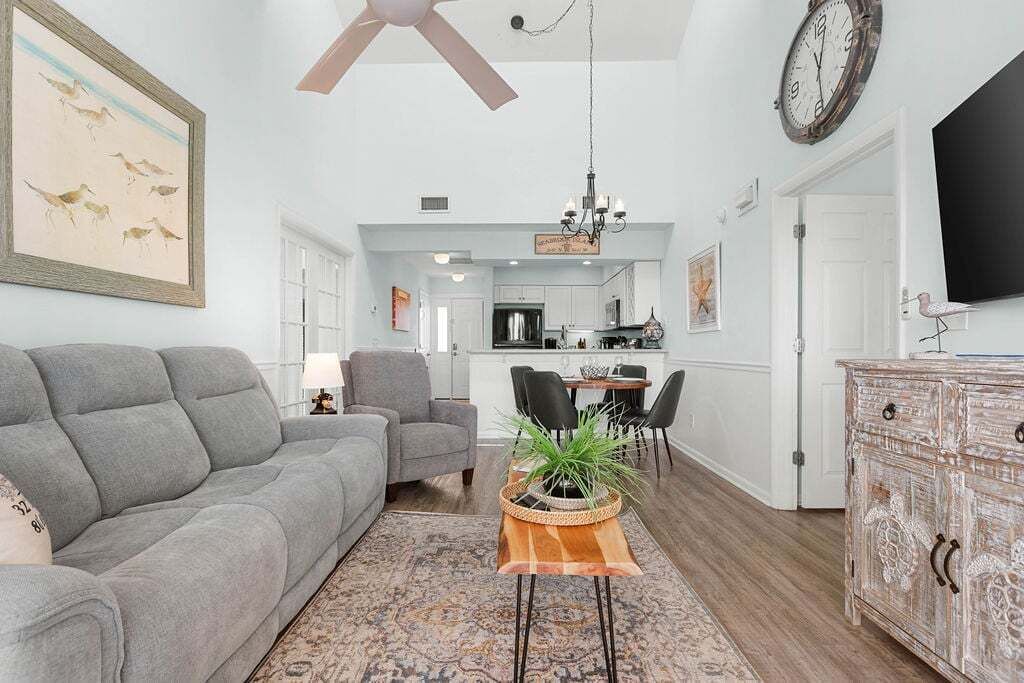 A living room with a grey sofa, a coffee table with a plant, a dining area with chairs, and a kitchen in the background.
