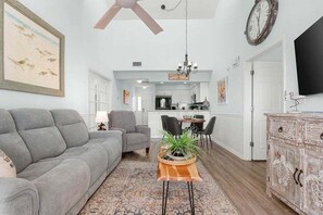 A living room with a grey sofa, a coffee table with a plant, a dining area with chairs, and a kitchen in the background.