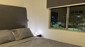 A neatly made bed with patterned bedding and two rolled towels, a bedside lamp, and a window with a view of greenery and buildings. - 5 minutes from the beach and pool (manzanillo)