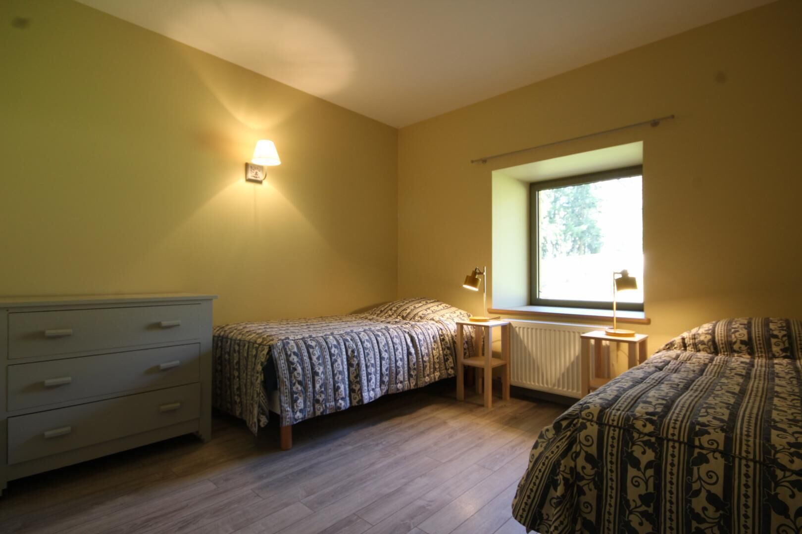 Une chambre avec deux lits, un bureau, une chaise, une commode et une fenêtre laissant entrer la lumière naturelle.