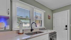 A kitchen with white cabinets, a stainless steel sink, and a dishwasher. There are two windows with blinds, a door with a keypad lock, and a small painting on the wall.