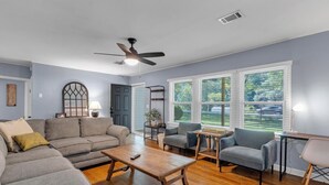 A living room with a gray sofa, a wooden coffee table, a gray armchair, and a small table with a lamp. There is a ceiling fan, a window with blinds, and a door.
