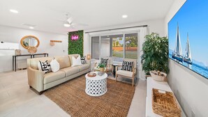 Un salón moderno con sofá, una mesa de centro y sillones, un ventanal con vista al mar, y un letrero de neón en la pared.