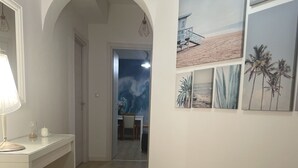 Käytävän lattia on puuta, siellä on valkoinen työpöytä, jossa on lamppu, ja seinällä on kehystettyjä kuvia rannasta ja palmuista.
