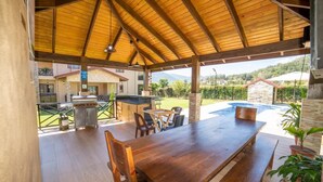An outdoor covered patio with a wooden table and chairs, a barbecue grill, and a view of a swimming pool and greenery.