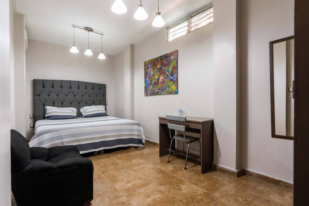 A bedroom with a bed, a desk, and a chair. There is a painting on the wall and a window with blinds.