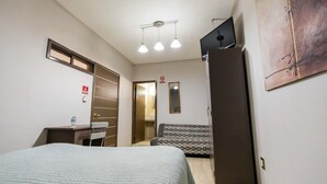 A compact hotel room with a bed, a desk with a chair, a sofa, a mirror, and a door leading to a bathroom. - Hotel Boutique 5ta avenida (Córdoba)