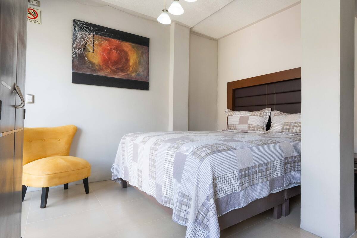 A bedroom with a bed, a chair, a painting, and a water bottle on a nightstand.