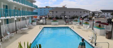 A swimming pool with a blue water feature, surrounded by lounge chairs and a building with balconies.