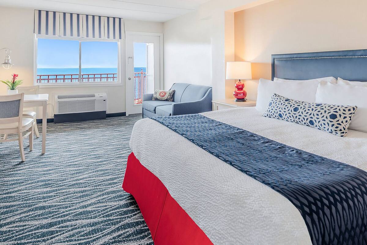 A hotel room with a bed, a desk, a chair, a window with a view of the sea, and a balcony.