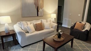 Ein Wohnzimmer mit einer Couch, zwei Beistelltischen, einem Couchtisch und einem Sessel. An der Wand hängt ein Gemälde und auf jedem Beistelltisch steht eine Lampe.