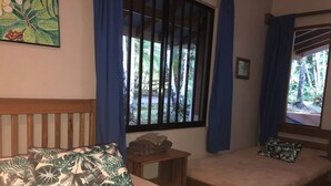 Ein Zimmer mit zwei Betten, eines mit Holzkopfende und das andere mit Holzrahmen. Es gibt ein Fenster mit blauen Vorhängen, einen kleinen Tisch mit einem Handtuch und ein an der Wand hängendes Bild.
