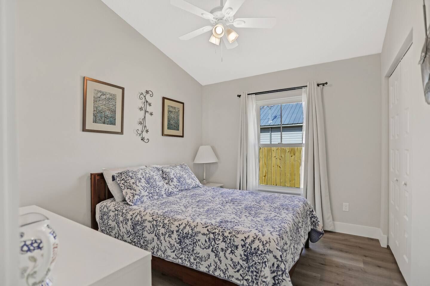 Une chambre à coucher avec un lit, un ventilateur de plafond, une fenêtre avec des rideaux et des tableaux encadrés accrochés au mur.
