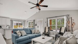 Un salon moderne avec un canapé bleu, une table basse blanche et un fauteuil en cuir noir. On y trouve un ventilateur de plafond, une fenêtre donnant sur un espace verdoyant, et un coin cuisine à l’arrière-plan.